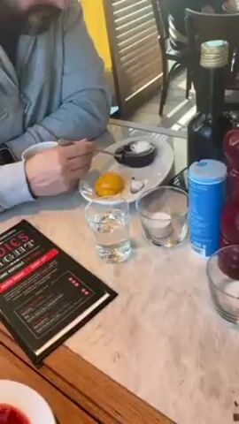 The video shows a table in what appears to be a restaurant. Initially, a hand holding a spoon is visible, reaching toward a chocolate dessert on a white plate. Beside it is a yellow fruit, possibly a peach or mango, and a small glass of water. A black menu and a bottle are also on the table. The video then focuses on a second dessert, a panna cotta with strawberry sauce and sliced strawberries, also on a white plate with a spoon. A phone is visible in the background.