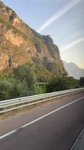 The video shows a view from a vehicle moving along a highway. The highway has two lanes separated by white lines and a white guardrail runs along the right side of the road. To the left of the highway is a steep, rocky mountain covered in greenery. Trees are visible at the base of the mountain. The sky is blue with some thin white clouds. In the distance, more mountains can be seen. The lighting suggests it is either early morning or late afternoon. The perspective indicates the camera is looking out from a side window of the moving vehicle. There are no people clearly visible.