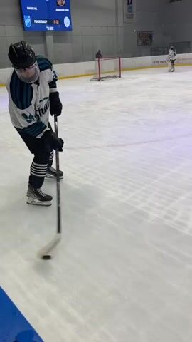 The video shows a person playing hockey on an indoor rink. The hockey player is wearing a white, green and black jersey and black pants and skates. The player has the puck, skates around the rink, and shoots it toward a net. There is a goalie in the net, wearing white pads and helmet. The rink is white with blue lines and borders. The background includes bleachers, walls, an electronic scoreboard, and other players. The building has a metal grid ceiling with blue lights.