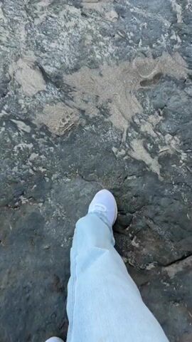 A first-person perspective video shows feet wearing white sneakers and light-colored pants walking on a rocky, sandy beach. The camera pans to reveal the ocean with waves crashing onto the shore and a clear blue sky.