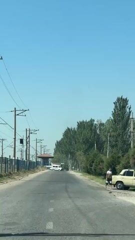 The video begins with a POV shot from a vehicle driving on a paved road. Overhead power lines stretch along the left side of the road, supported by poles. A fence runs parallel to the power lines. In the distance, a small building with a red roof is visible, with a white van parked in front of it. A line of tall green trees forms a border along the right side of the road, with some smaller trees and bushes closer to the roadside. A person is standing on the right side of the road, near a yellow vehicle, facing away from the camera. The sky is clear and blue. The vehicle continues to drive down the road, passing by the trees and the person on the right. A faint contrail is visible in the sky.