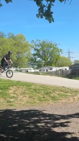 A person on a BMX bike rides onto a BMX track with multiple hills and jumps. Other people are also riding on the track. The day is sunny with a clear blue sky and green trees. The camera follows the person on the bike as they navigate the track.