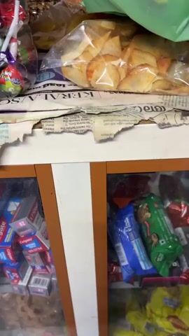 The video opens with a close-up shot inside a small shop or snack stall in Chithirapuram, India. On a counter, various packaged snacks are displayed, including what appear to be chips or fried items in clear plastic bags, resting on torn newspaper. A bright green plastic carry bag sits on top of these snacks, and through its translucent material, a package with the visible text "FINEST POTATOES" can be discerned. Below the counter, shelves hold more packaged goods like chocolate bars and other snacks.

From the 00:01 mark, the focus shifts to an elderly man with a grey beard and short grey hair, wearing a dark-colored hooded jacket. He is positioned behind the counter, looking calmly towards the camera. Behind him, shelves are stocked with numerous small, brightly colored snack packets hanging, indicating a variety of offerings. More packaged snacks are also visible on the counter beside him. The scene is lit by indoor lighting, depicting a typical local convenience or snack store environment.