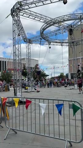 The video shows a group of people performing on a large metal structure in a public square. The structure consists of towers, interconnected by beams and ropes. Several people are climbing up the towers, while one person is swinging from a rope between the towers.  A crowd of people is watching the performance from behind a gray metal fence, which is decorated with colorful triangular flags. There are buildings visible in the background, and the sky is partly cloudy.