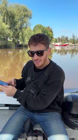 The video shows a man in sunglasses taking a photo of a brown river with his phone. He's wearing a black sweatshirt, blue jeans, and a small black bag across his chest. He's sitting on the side of a white boat. The camera then pans to show a large bottle of water on the boat, and the river flowing between lush green trees and vegetation. The water is opaque and brown. The sky is blue.