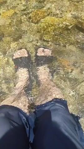 The video shows a first-person perspective of someone's feet submerged in clear, shallow water. The feet are wearing black sandals. The water is flowing gently over a rocky bottom, with some green moss or algae visible on the rocks. The person is wearing dark pants that are wet from the water. The sun appears to be shining, casting light and reflections on the water's surface. The camera perspective is looking down at the feet.
