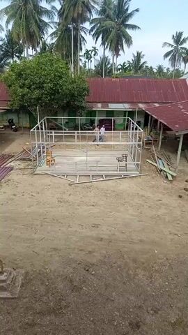 Here's a detailed description of the video:

The video shows a construction site in what appears to be a tropical area. A metal frame structure is being built on a sandy lot. The frame is rectangular and seems to be the skeleton of a small building.

A person wearing a white shirt and dark pants walks around the frame structure. The person appears to be observing or checking the progress of the construction.

In the background, there are several houses with red roofs and green trim. Palm trees are abundant, surrounding the construction area. There are also pieces of wood and metal sheeting scattered around the construction site.