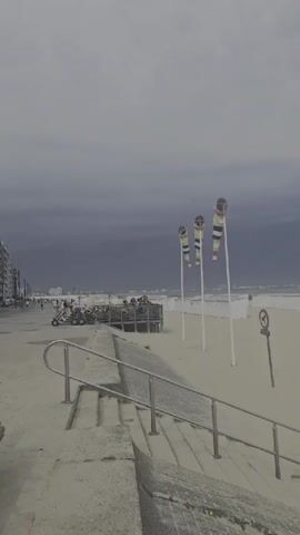 The video shows a beach scene under an overcast sky. In the foreground, a concrete embankment with a metal railing leads down to a sandy beach. Further back, several colorful windsock-like flags are planted in the sand. A few small white beach huts are visible in the distance. On the sand, two people are walking away from the camera. The sea is visible in the background.