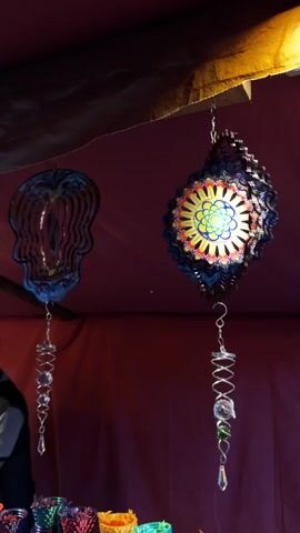Two metal wind spinners hang from a wooden beam, their designs a skull and a floral pattern respectively. The skull is painted in a Dia de los Muertos style, while the flower is painted in yellow, blue, and red. Each spinner has a silver chain attached, ending with a crystal-like decoration. Below them, various colorful containers sit on a table. A person, partially visible on the left, appears to be standing behind the table. The background features a red curtain-like fabric.