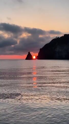 The video captures a serene sunset over the sea in Lipari, Italy. The sun, a vivid orange-red orb, is seen descending between two dark, silhouetted rocky formations or islands in the distance. The sky above is partly cloudy, with dark grey clouds contrasting with the warm hues of the sunset. A prominent, shimmering reflection of the sun stretches across the calm water towards the foreground, creating a path of intense orange and red light. As the video progresses, it slowly zooms in on the setting sun, making its fiery color and the silhouettes of the rocks more pronounced. The gentle sound of water or waves can be heard, adding to the tranquil atmosphere of the scene. The overall impression is one of natural beauty and peacefulness as day transitions to night.