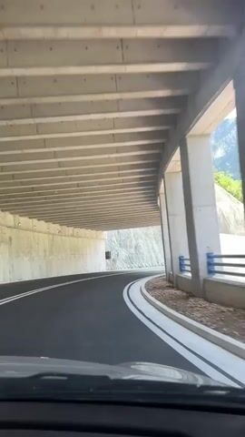 The video captures a first-person perspective of a car driving on a paved road. The road curves to the right and goes under a concrete overpass supported by columns. The overpass ceiling consists of parallel concrete beams. To the left of the road, a steep, rocky mountainside is visible, partially reinforced with concrete structures. To the right, there is a metal guardrail with blue and white elements, separating the road from a dirt embankment with some vegetation and trees. As the car emerges from under the overpass, the view opens up to a scenic mountainous landscape with lush green slopes and tall trees under a bright blue sky with scattered white clouds. The road has clear white lane markings.