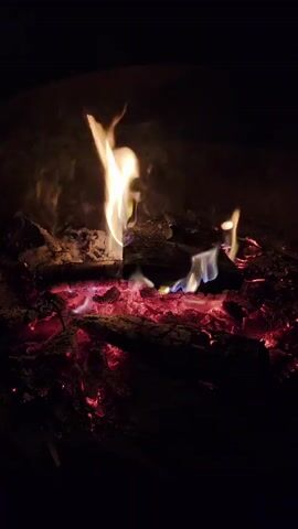 A close-up shot shows a campfire with glowing red embers and flickering flames against a dark background. The fire is composed of logs, ash, and burning wood. The flames are primarily yellow and white, with some hints of blue and purple at the base. The smoke is light and wispy, blending into the darkness. The overall impression is one of warmth and intensity, with the fire being the sole source of light and color in the frame.