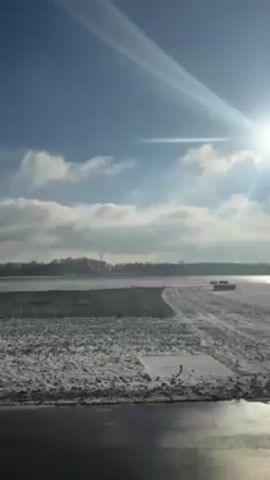 The video shows a bright, sunny day at what appears to be a partially snow-covered area, possibly near a body of water. The sun is high in the sky, casting rays downwards. There are scattered clouds. In the distance, trees line the horizon. In the foreground, the ground is a mix of dark pavement or soil and patches of snow. The overall scene is cold and bright.
