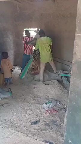 The media file shows an indoor scene with several people and objects. The setting appears to be a rustic, unfinished building with dirt floors and rough walls, possibly a workshop or storage area. Sunlight streams in from an opening in the wall, suggesting it is daytime.

The subjects include at least three children and two adults. One adult, wearing a striped shirt, stands near a machine. Another adult, in a light green shirt, is also operating or attending to the machine. The children are positioned around the adults and the machine. One child, wearing a light brown shirt, walks towards the front of the frame, while another, in a blue garment, stands to the left. A third child's head is visible in the foreground, suggesting they are close to the camera.

The central activity appears to be the operation of a machine, possibly for grinding or processing materials. Sacks of what appears to be grain or powder are visible on the floor, along with some scattered debris. The overall atmosphere seems industrious, with people engaged in a task. The lighting suggests it is daytime, and the simple construction of the building might indicate a rural or developing area. No specific emotions are clearly discernible from the still images. There is a partial view of text on a gray sack that reads "SACK".