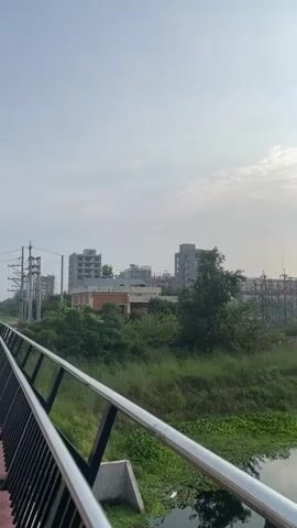 The video pans across a scene of a river and some construction sites. The river is visible in the foreground, with some plants growing along its banks. In the background, there are several unfinished buildings, some with exposed rebar. There are also electrical towers and power lines visible. The sky is cloudy with some sunlight breaking through. The overall impression is one of urban development in a natural setting.