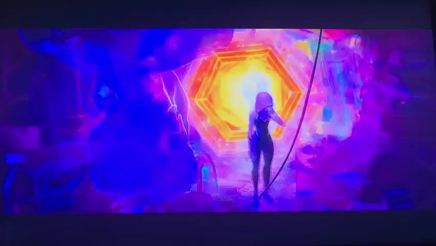 The media file depicts a scene from an animated film, likely a superhero movie given the character's pose and the vibrant, surreal environment. A solitary female figure, silhouetted against a powerful light source, stands at the center of the frame. She appears to be wearing a sleek, form-fitting outfit, possibly a costume. Her posture suggests she is in the midst of an action or a moment of emotional intensity, perhaps reacting to the overwhelming visual display around her.

The setting is highly stylized and abstract, dominated by a large, luminous, hexagonal portal or energy source radiating intense yellow and orange light. Surrounding this central element are chaotic, fragmented shapes and a riot of colors, including deep blues, purples, and pinks. These elements create a sense of otherworldly dynamism and perhaps interdimensional travel or a reality-bending event. The overall atmosphere is one of immense energy, spectacle, and a touch of disorientation, possibly evoking awe, fear, or determination in the character. The environment feels like a constructed, digital, or a highly fantastical space rather than a natural landscape. There is no discernible text within the visible frame. The time of day and weather are irrelevant to this artificial, visually driven scene. The implied emotion is one of intense focus and perhaps a struggle or significant moment for the character.