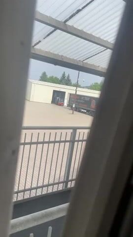 This media file is a short, grainy video clip filmed through a slatted barrier, likely a fence or window frame. The perspective is from an elevated position looking down and out towards an outdoor industrial or commercial area.

The primary subjects are a building with large bay doors, a black semi-trailer truck parked in front of it, and a small red forklift. The building appears to be a warehouse or distribution center, suggested by its functional design and the presence of the truck and forklift. The truck has lettering on its side, partially visible, that includes "DC TRANS" and "TIGERS" in orange text. There is also some smaller text below it, but it is too blurry to read. A security camera is visible mounted on a pole near the truck.

The scene depicts a typical daytime operation at a logistics or transport hub. There are no people clearly visible in the frame, suggesting the activity may be paused or taking place inside the building. The ground is paved with interlocking bricks or pavers. In the background, there are several evergreen trees and a cloudy sky, indicating overcast weather. The overall atmosphere is neutral, functional, and somewhat industrial. The slatted foreground creates a sense of distance and observation, as if the viewer is looking in from the outside. The location context of Teplice, Czechia, aligns with the typical European industrial landscape.