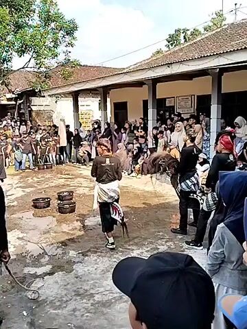The video depicts an outdoor cultural performance, possibly a traditional dance or reenactment. A crowd of spectators, some masked, are gathered on the right and in the background, observing the event. The performance area appears to be a courtyard with a dirt and water surface. In the foreground, a man in a black shirt and white shorts stands facing away from the camera, holding a rope-like object. To his right, a performer dressed in traditional attire, including a striped sarong and head covering, is engaged in the performance. This performer is interacting with what appears to be a prop, possibly a stylized horse or animal figure made of straw or similar material, which is on the ground. The performer is in motion, bending and possibly manipulating the prop. In the background, other performers, also in traditional clothing, are standing. There are also several round, dark objects, possibly drums or containers, arranged in a line on the ground. The environment includes buildings with tiled roofs and trees. The time of day appears to be daytime, with clear skies.
