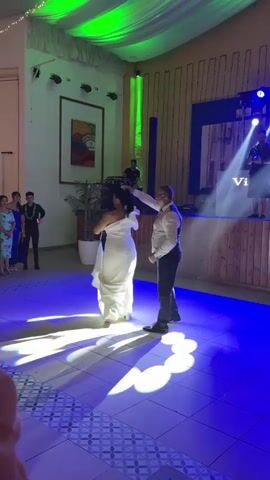 The video shows a man and a woman dancing on a tiled floor in a dimly lit venue. The woman is wearing a long, white dress with ruffled sleeves and the man is wearing a white dress shirt, a dark vest, and dark pants. They are holding hands and dancing in a circle, with the woman's dress flowing around her. Blue and green lights illuminate the dance floor, casting shadows. In the background, there are people standing and watching, a DJ setup with a keyboard and speakers, and a stage with lights and a banner that appears to read "Vicent 2023". The environment appears to be a banquet hall or event space.