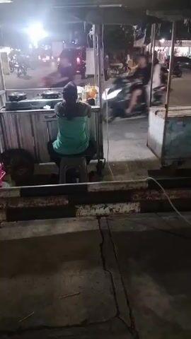 A person sits on a stool facing away from the camera, behind a food cart. The food cart has a metal exterior and a canvas covering. The person is wearing a teal shirt and dark pants, with their hair in a braid. They appear to be resting or waiting. The background shows a street scene at night, with traffic and streetlights visible. Several motorcycles are passing by, their headlights illuminating the area. The overall environment appears to be an urban street at dusk or night.