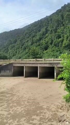 The video pans across a beach with a river running into it. There are hills covered in lush green vegetation in the background. The sky is cloudy and overcast. The beach is sandy with some debris scattered around. There are some small structures along the coastline, including what appears to be a covered seating area and possibly a small hut. A road runs behind these structures. A concrete bridge with three archways spans the river. The water in the river appears murky and shallow. The waves are relatively small and gentle. There are tire tracks and footprints visible in the sand.