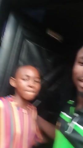 The video shows two individuals, a young person and an adult, standing in front of a dark, textured surface, possibly a door or a wall. The young person on the left is wearing a brightly colored striped shirt with vertical bands of purple, pink, yellow, blue, and green. They are looking towards the camera with an open mouth, as if speaking or singing. The adult on the right is wearing a black polo shirt with green stripes. They are also looking at the camera, smiling and gesturing with their hands. The lighting is somewhat dim, with the faces of the individuals illuminated. In the background, above the adult's head, there is a small rectangular sign with text that is difficult to read due to the resolution and angle, but it appears to have a picture on it. The overall impression is of a casual, informal interaction.