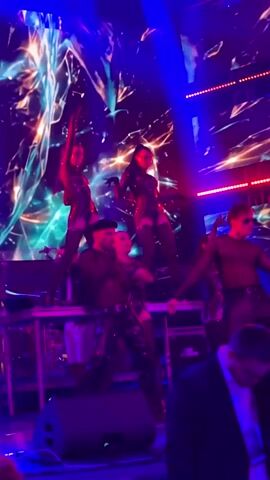The video shows a group of performers on a stage with a large screen displaying vibrant, abstract, and colorful visuals. There are five performers visible on stage, some of whom are positioned on elevated platforms. Two female performers in shiny, dark, form-fitting outfits, possibly with cutouts, are standing on higher platforms, with their arms extended to the sides, in dynamic poses. Below them, three other performers are on a lower level. One male performer in a dark top and pants is on the left, reaching out. Another male performer in a dark top and sunglasses is on the right. A female performer in a similar outfit to the ones above is in the center, also striking a pose. The stage is lit with bright blue and red lights. In the foreground, a man in a black suit jacket and white shirt is partially visible, looking towards the stage. The overall atmosphere appears to be that of a live performance, possibly a concert or show, with a focus on visual effects and choreographed movement.