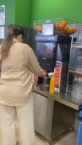 The video depicts a woman in a tan shirt and white pants, standing at an automatic orange juicing machine. She feeds oranges into the machine, and orange juice pours into a plastic bottle placed under the dispenser. The machine, branded Zummo, has a basket on top filled with oranges and a digital display showing a price of 109.00. To the right of the machine are empty plastic bottles on a metal rack. The backdrop is a green wall. The machine has a stainless steel base with a cupboard and is on wheels. A sign next to the dispenser shows instructions on how to use the machine.