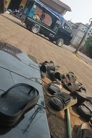 The video shows an outdoor scene with a dirt ground and a tiled surface in the foreground. A collection of black sandals are arranged on the dirt, some in a line and others clustered together. Some of the sandals appear to be brand names like Puma. Behind the sandals, a small truck or utility vehicle is parked on a paved area. The truck has a blue canopy and appears to be carrying items. To the left of the truck, there is a small building with a brown door and a window. The background shows trees and a building under construction or repair. The lighting suggests it is daytime.