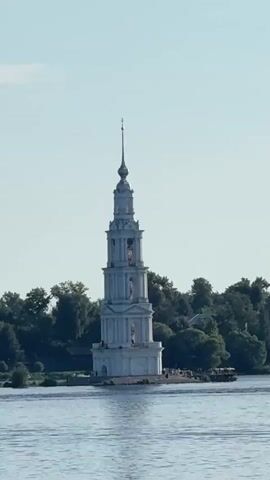 The video shows a tall, white bell tower standing in the middle of a body of water. The bell tower is multi-tiered and topped with a spire. Trees and buildings are visible on the shore in the background. The water is calm and reflects the sky, which is a light blue with a few scattered clouds. A person is seen paddling a stand-up paddleboard on the water in the foreground, moving away from the bell tower. The scene appears to be tranquil and scenic, likely during daylight hours.
