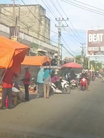 A busy street scene unfolds with several people and motorcycles. On the left, a man in a red shirt stands near a market stall with an orange awning. Two women wearing hijabs, one in light blue and the other in grey, stand together near the stall. A white motorcycle with a red seat is parked in front of a stall covered by a red umbrella. Another white motorcycle is to its right, with a person wearing a purple hijab on it. A person wearing a red hijab is on a red motorcycle to the left of the white motorcycle. In the background, a large billboard advertising "BEAT KRETEK" is visible, along with power lines and buildings. More people and motorcycles are scattered along the street, suggesting a market or a public gathering. The ground is paved, and there appears to be some debris on the side of the road. The overall impression is a lively, active street environment.