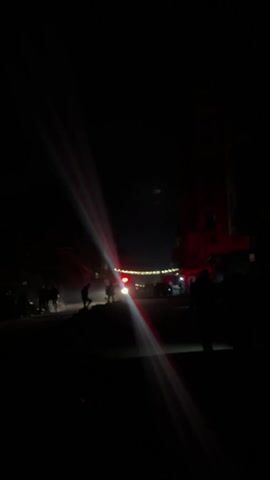 The video shows a street at night with an ambulance approaching. The headlights and red lights of the ambulance are visible, creating bright streaks of light in the dark. There are indistinct figures of people on the sides of the street. The background includes some buildings and string lights. The overall scene is dark and blurry. The sound of an ambulance siren is audible.