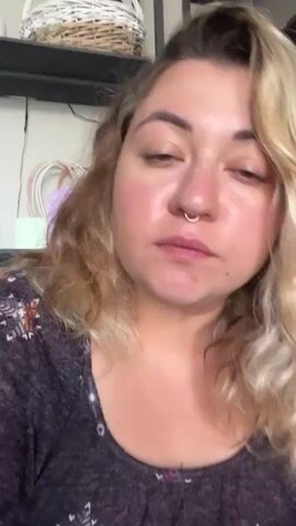 A young woman with blonde, wavy hair and a nose ring is looking at the camera. She has rosy cheeks and is wearing a dark-colored shirt with a pattern. The background appears to be a room with shelves containing baskets and other items. The woman is speaking and gesturing with her hands.