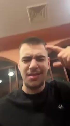 The video shows a young man filming himself and his friends at what seems to be a restaurant or bar. He starts with a close-up, points to his forehead, then gestures downwards to include a young woman with long, curly hair sitting next to him. She is also facing the camera, smiling and singing along to the music. Another young man with glasses is visible in the background. The first man then puts his arm around the woman, and she smiles at the camera. The background shows typical restaurant decor with tables, chairs, and decorative elements, suggesting a relaxed and social environment.