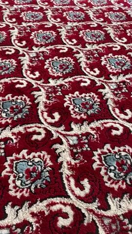 The video shows a close-up, low-angle, panning shot of a red carpet with a repeating pattern of cream and light blue floral and paisley designs. The camera moves from left to right, revealing more of the carpet's texture and pattern. The lighting is consistent throughout, suggesting an indoor environment with ambient light.