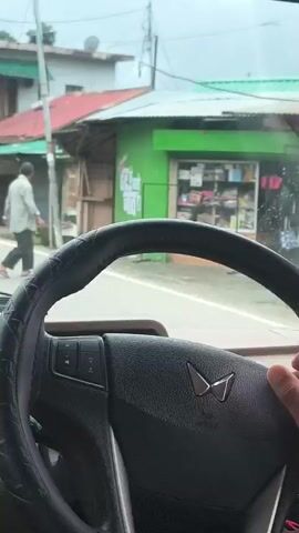 The video shows a person driving a vehicle. Their hands are on the steering wheel, and they are wearing a light blue shirt and a yellow bracelet. The interior of the car is visible, including the dashboard, steering wheel, and gear shift. Outside the car, there is a street with buildings and other vehicles. The weather appears to be cloudy or rainy, as evidenced by reflections on the car's windows and the general lighting. The driver is focused on the road ahead.