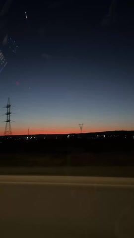 The view from inside a vehicle shows a vibrant sunset in the distance, with a gradient of red fading into a dark blue sky. Power lines and transmission towers stand silhouetted against the colorful horizon. The vehicle is moving along a road or highway, with a dividing barrier visible in the foreground. Another car passes by in the opposite direction. The interior of the vehicle is visible through reflections in the windshield, and the side mirror is also visible.