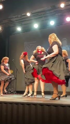 A group of five women in matching black shirts and skirts with ruffled underskirts perform a choreographed dance on a stage in front of a large screen. The women are all facing away from the camera, with their backs mostly to the audience. The skirts are voluminous and appear to be made of a light material, as they are swirling around the dancers' legs. Some of the women have red hair accessories, and one has a red scarf around her neck. The stage is lit by spotlights, and there is a microphone stand on the left side of the stage. The screen behind the dancers displays a logo and text that is partially obscured. The overall impression is a lively and energetic performance.