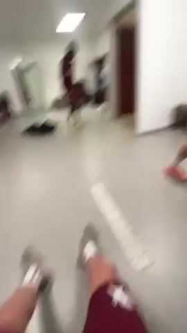 The camera pans across the interior of a locker room. Several people wearing maroon and white clothing are seated. The floor is gray tile. There is a brown wooden bench. There are several bags and articles of clothing lying on the floor. The room is illuminated by fluorescent lighting.