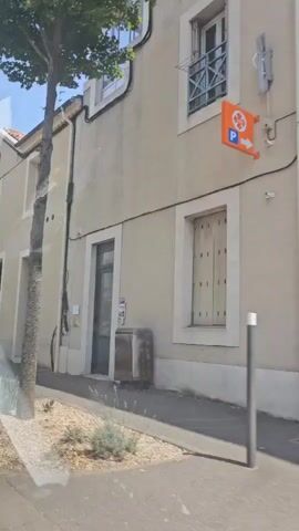 The video shows a street view of buildings. On the left, there is a building with a sign that says "OPTIMES LASSERTE". Further down the street, there is another building with a sign that says "SAMSIC EMPLOI". On the right side of the street, there is a tree and a building with a sign that says "L'IMMOBILIER EN GASCOGNE". The sky is blue and it appears to be a sunny day. The camera is moving, giving the impression of driving past the buildings. There are no people visible in the video.