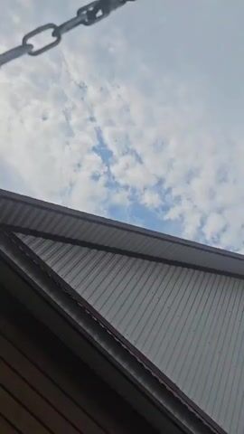 The video shows the roofline of a building made of light gray corrugated siding with dark brown trim. Above the building, a blue sky is visible with scattered white clouds. A small airplane is visible in the distance, flying across the sky. A chain dangles in the corner of the frame.