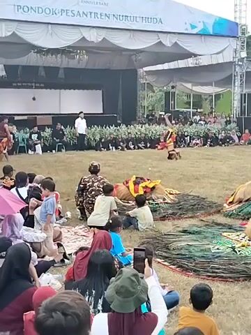 The video captures an outdoor cultural performance at "Pondok Pesantren Nurul Huda" in Plosorejo, Indonesia, celebrating its 41st anniversary. The scene is set during the day under a large white tent that provides shade for a stage. In the foreground, a diverse audience, including many children, is seated on the grass, some holding umbrellas for shade. A performer in traditional attire, possibly a mask or costume with a horse-like effigy and elaborate headwear, is actively engaged in a dance or ritual in the middle of a grassy field. Behind the performer, a seated group of men, likely participants or organizers, are dressed in black and white attire. Further back, a larger seated audience is gathered in rows on the grass, facing the stage. The atmosphere appears festive and engaging, with the audience attentively watching the performance. The event is taking place in a rural or semi-rural setting, indicated by the open grassy area and the presence of trees in the background. The sky is overcast, suggesting it might be a cloudy day. The overall impression is of a community gathering celebrating a significant milestone for their pesantren.