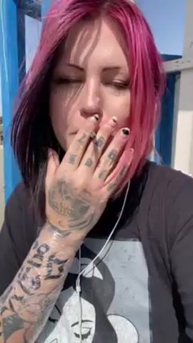 A person with pink and black hair, tattoos covering their left arm and the back of their left hand, is smoking a cigarette. The person, wearing a black t-shirt and white earphones, exhales smoke into the air. They are standing outdoors, with blue and white structures visible in the background. The sky is light blue.