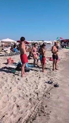The video shows a beach scene on a sunny day with a clear blue sky. In the foreground, on the sandy beach, are four people. From left to right, a man wearing red swim trunks stands facing left. Next to him, a woman in a bikini stands facing right. Beside her, a boy in red swim trunks stands facing right. Further to the right, a man in black swim trunks stands facing right. There are beach umbrellas and chairs set up further back on the beach, with many people sunbathing and swimming in the turquoise water. Some people are walking along the shoreline, and children are playing in the water. The waves are gently lapping at the shore.