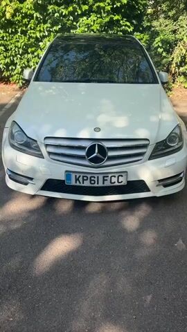 This is a vertical video of the front of a white Mercedes-Benz sedan parked outdoors on an asphalt surface. The car is centered in the frame. The car has a black roof and a blue and white license plate that reads "KP61 FCC." The car is facing the camera, and the front grille and headlights are clearly visible. Behind the car, there is a dense green hedge or vine-covered wall. Sunlight is casting dappled shadows on the ground and the car. The video is static with no movement.