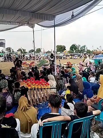 The video shows a crowded outdoor event under a large white tent. A group of people, many wearing traditional clothing and headdresses, are seated on a blue tarp in the foreground, facing a performance area. Some are playing musical instruments, including what appears to be a gamelan. Other attendees are seated on chairs or on the ground behind the tarp, watching the performance. In the background, more tents and people are visible, suggesting a larger festival or gathering. The ground appears to be grass.