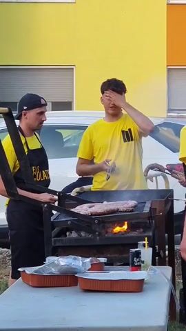 This video captures a lively outdoor gathering, likely a barbecue or outdoor meal, in Sant Joan Les Fonts, Spain. The scene is set against the backdrop of a vibrant yellow building with visible windows and what appears to be a white car parked behind the subjects. The time of day seems to be daytime, with ample natural light.

The primary subjects are two young men. The man on the left is wearing a black baseball cap and a black apron over a yellow t-shirt. His apron has text that is partially visible, reading "ELS COLATS". He is holding a pair of tongs, actively engaged with the grill. The man on the right is wearing a bright yellow t-shirt with a black graphic design on the front. He is holding a drink in his left hand and appears to be touching his head with his right hand, possibly indicating warmth or a casual interaction.

The central focus of the activity is a barbecue grill, with flames visible underneath a rack holding several large sausages. The sausages are in the process of being cooked. In the foreground, a white table holds several foil food trays and a can of "Estrella Galicia", a Spanish beer, along with a glass of a yellow drink. Another person's hand can be seen in the background holding tongs, also attending to the grill.

The overall atmosphere appears relaxed and informal, suggesting a social gathering or celebration. The bright colors of the building and clothing, along with the activity of grilling, contribute to a sense of warmth and enjoyment.