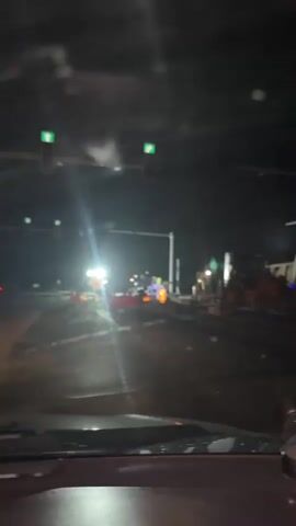 The video captures a nighttime scene from inside a vehicle, looking out through the windshield at a construction zone. The time is night, with artificial lights illuminating the area, causing significant glare on the wet or reflective road surface.

In the foreground, two traffic lights are visible; initially, they display green arrows, then transition to orange/yellow lights. Ahead, bright work lights from construction equipment or portable setups illuminate the road. A prominent yellow diamond-shaped sign with a "men at work" symbol is visible, indicating active construction. Red and white plastic barriers, along with orange traffic cones, delineate the work area. Several pieces of heavy machinery, including what appears to be a mini excavator or skid steer loader, are parked or in use in the background. One person, likely a construction worker, is visible near the yellow sign. The scene suggests ongoing roadwork or maintenance during the night. The vehicle appears to be stopped at an intersection within or adjacent to the construction area.