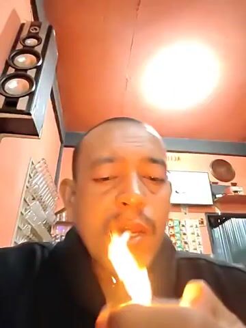 A man is lighting a cigarette in what appears to be an indoor establishment. He has a shaved head and a mustache, and he’s wearing a dark-colored shirt. He is looking directly at the camera. The room has a reddish-orange ceiling and a white light fixture. A tall speaker with three circular components is visible on the left wall. There is a rack with clear vials behind him. A sign and a TV are visible in the background. The man takes a drag from the cigarette as smoke fills the frame.