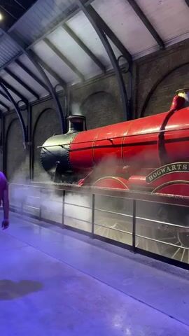 The media file shows the Hogwarts Express train, a replica of the train from the Harry Potter films, at a station. The train is bright red with black accents and emits steam. The words "HOGWARTS CASTLE" are visible on the side of the train, along with the number "5972". Two women are standing in front of the train, appearing to be tourists. One woman is wearing a denim skirt and black top, and the other is wearing a blue and white patterned top and shorts. They are looking at the train. The station appears to be an indoor setting with exposed brick walls and arched ceilings, suggesting a historical or themed location. The lighting is somewhat dim, with blue and purple hues cast on the ground, potentially indicating an artificial lighting setup for atmosphere. The overall impression is that of a popular tourist attraction, likely related to the Harry Potter franchise, with visitors admiring a key prop from the series. There is no information about the time of day or weather as the scene is indoors. The emotions of the people are not explicitly clear but appear to be casual observation and enjoyment.